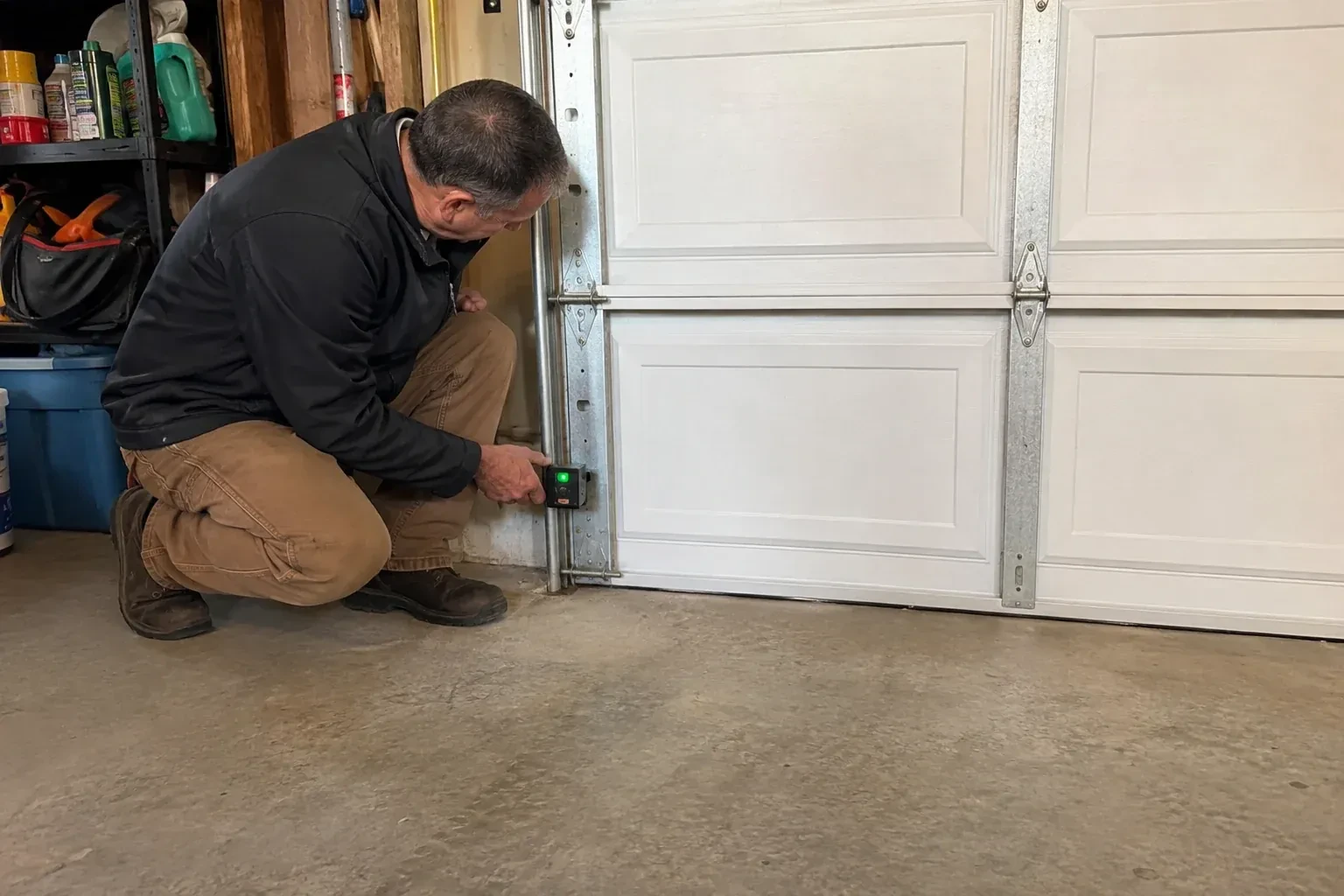 Technician kneeling beside a white paneled garage door, holding a small black box with a green light against the vertical track. — Photo-Eye Sensor Alignment or Replacement, Hamilton