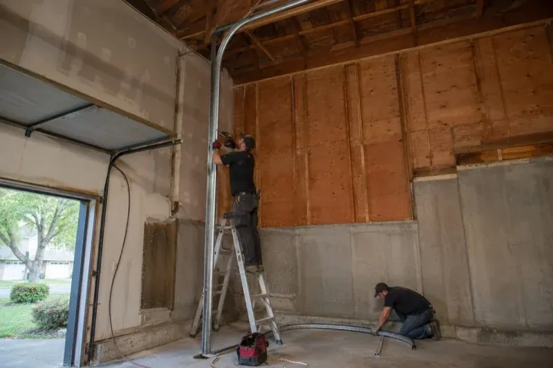 One person on a stepladder working beside a tall curved garage-door track while another kneels beside loose track sections in an unfinished garage. — High-Lift Garage Door Conversion, Hamilton